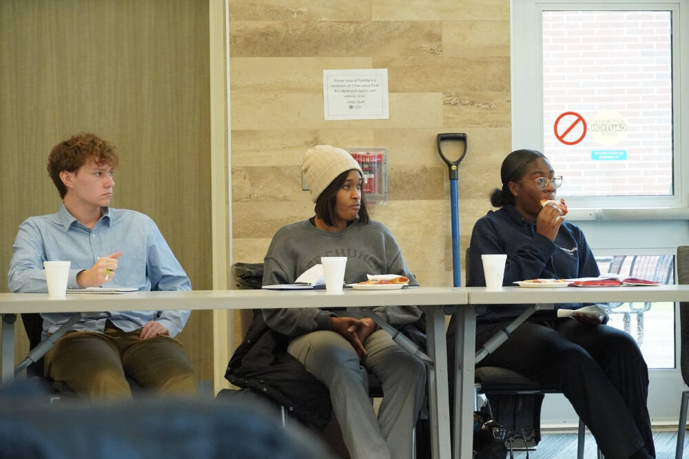 Three people sit at a long table with notebooks, paper cups, and plates of food in front of them. Behind them is a beige wall, a blue-handled shovel, and a door with a "no entry" sign. The setting appears to be a casual meeting or discussion.